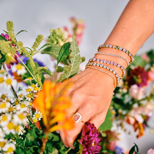 Hånd med Pure tennisarmbånd i guld blandt farverige blomster